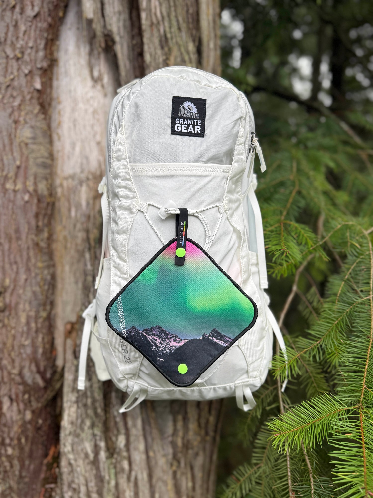 white backpack with a colorful pee cloth, leaning against a tree with greenery in the background