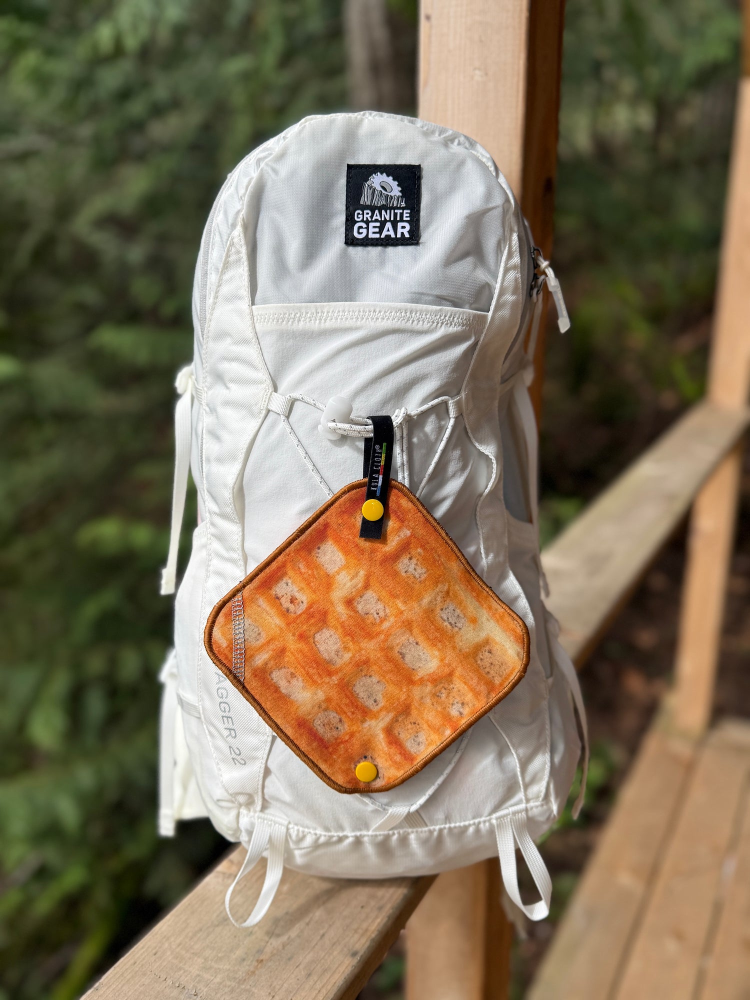White backpack with a waffle patterned pee cloth attached to it, featuring Granite Gear logo, on a wooden surface with greenery in the background.