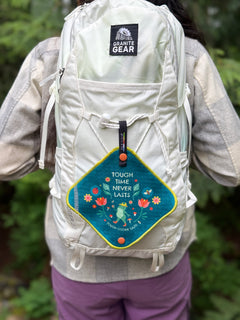 A person outdoors hiking with a pee green pee cloth on their white backpack. 