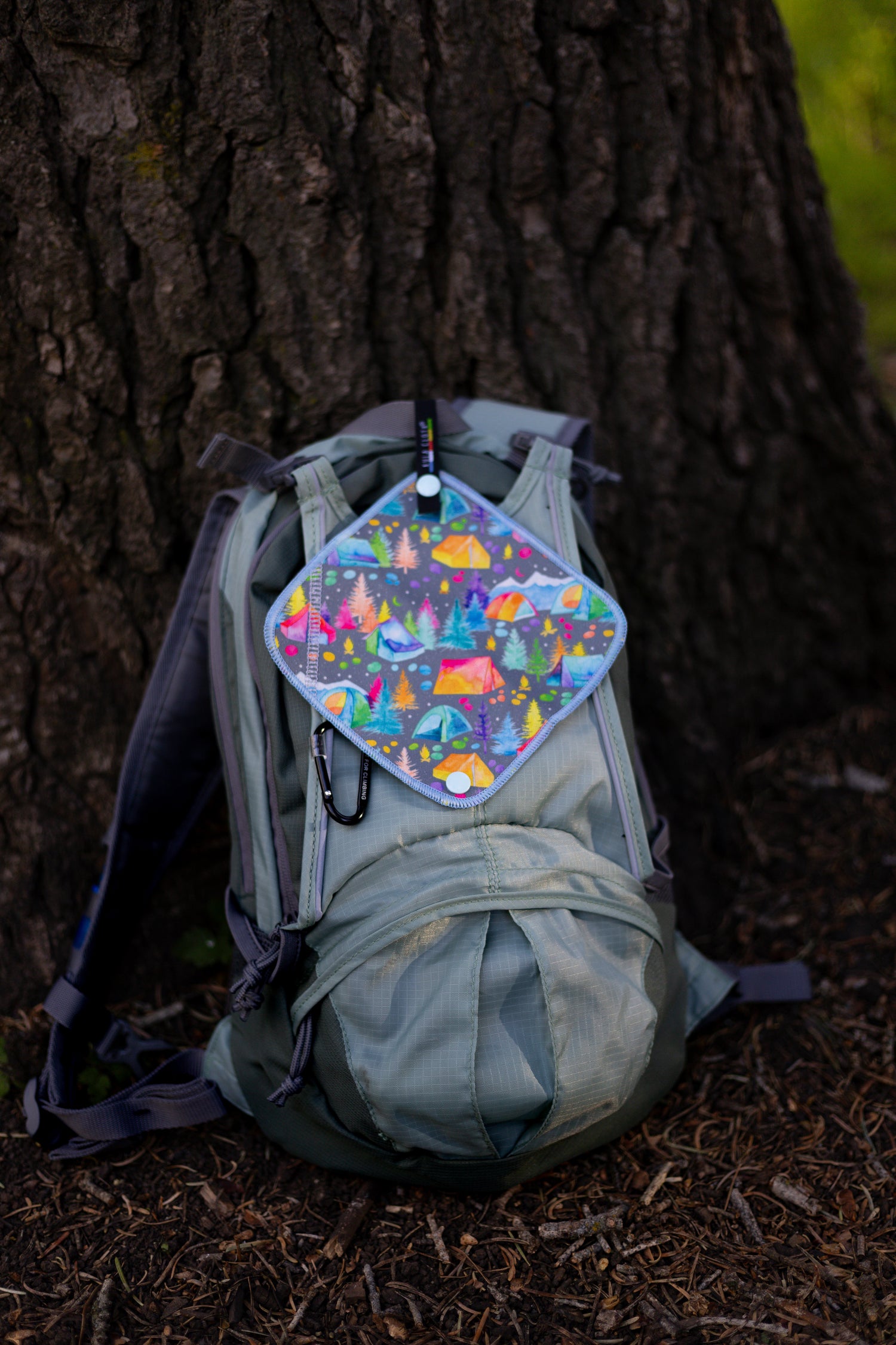 A sage green hiking backpack resting against a large tree trunk outdoors, with a colorful, patterned diamond-shaped rain fly attached to the front pocket.
